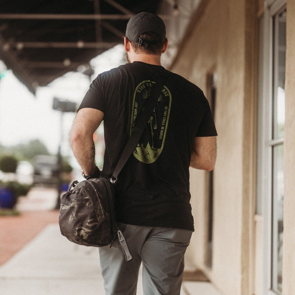 Photography Unknown Artist a man wearing a black t-shirt with a green and olive graphic design walks down a city sidewalk past a building with visible architectural details carrying a camouflage-patterned bag