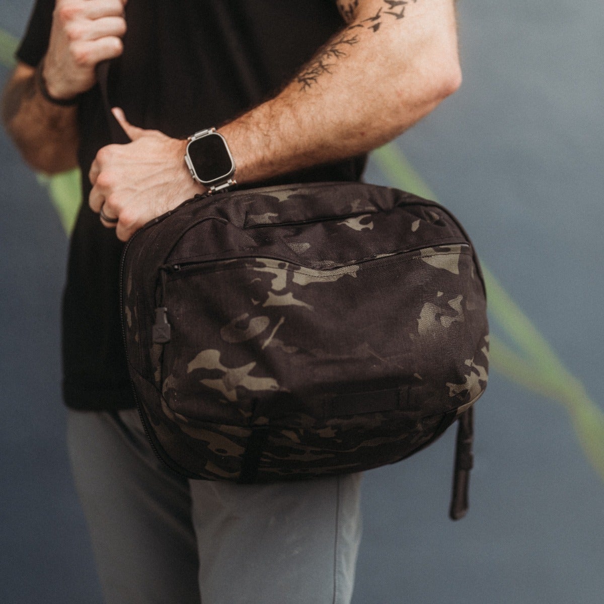 Photograph of a person carrying a dark camouflage-patterned bag with a silver watch and a subtle tattoo against a muted blue background with hints of green