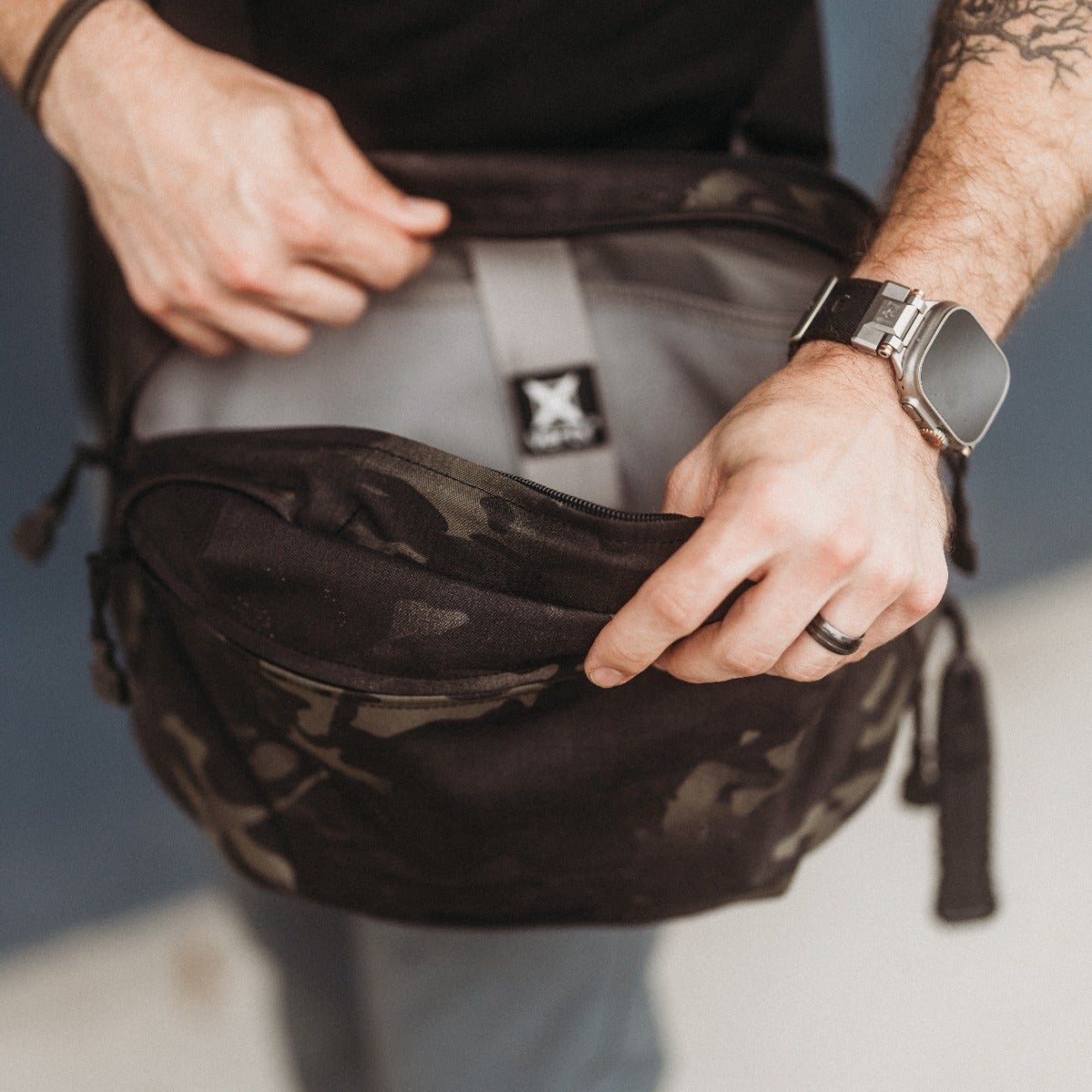 Photograph of a person displaying a dark camouflage-patterned bag with a grey interior and a branded tag, showcasing a wristwatch and a ring on their fingers.