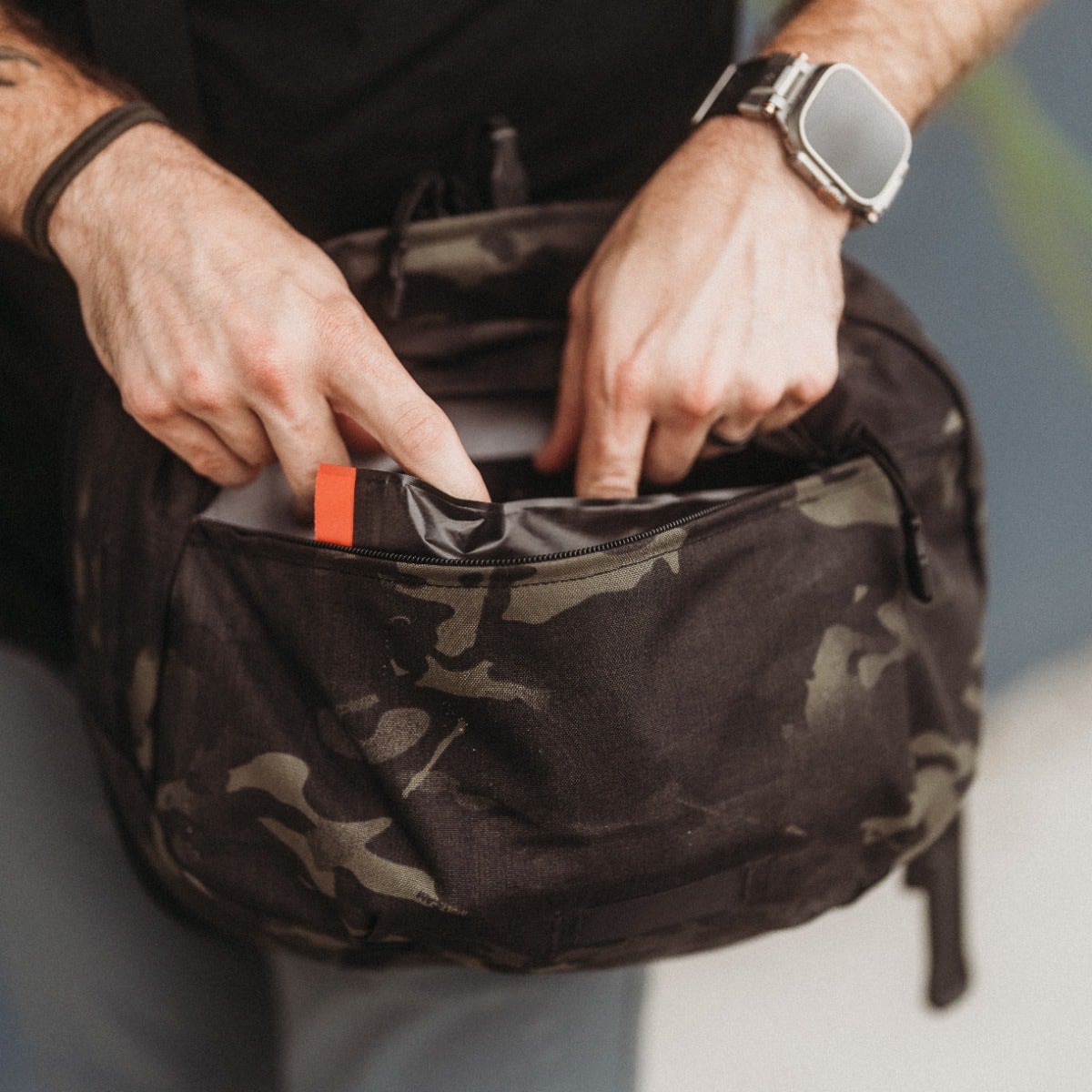 A photograph shows a person's hands unpacking a black and green camouflage backpack with a red zipper pull and a dark gray waterproof inner liner