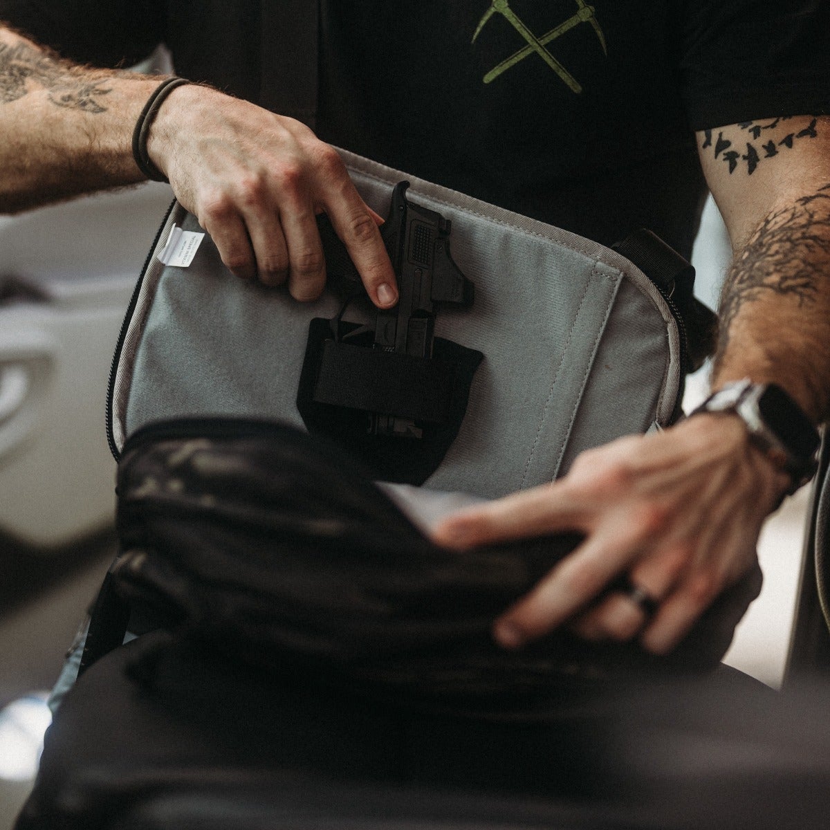 Photograph of a person's hands placing a handgun into a gray bag with a black strap featuring a dark gray backpack a black ring and tattooed arms