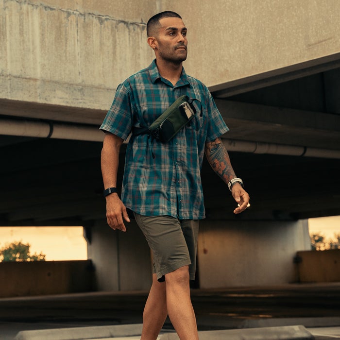 Photograph of a man wearing a teal plaid shirt, olive shorts, and a small green fanny pack walking under a concrete overpass with visible pipes and a sunset glow.