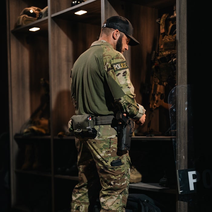 Photograph Police officer in a dimly lit armory preparing equipment near camouflage uniforms and riot gear a black pistol in a holster and a dark green POLICE patch are visible