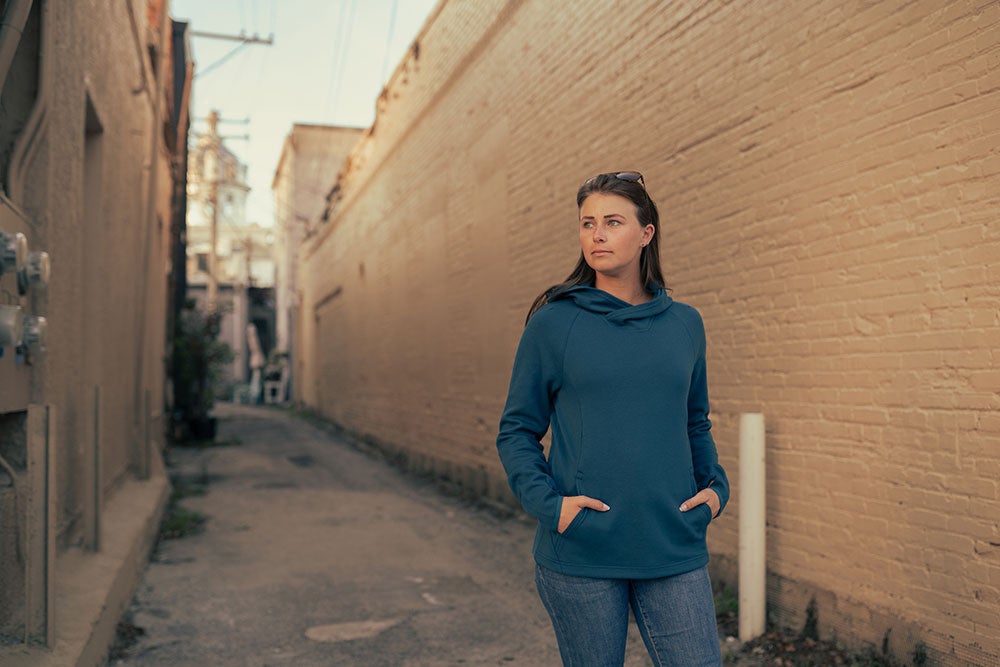 Photograph of a woman in a teal hoodie and jeans standing in a brick alleyway with a visible building in the distance featuring warm-toned brick walls and subtle greenery