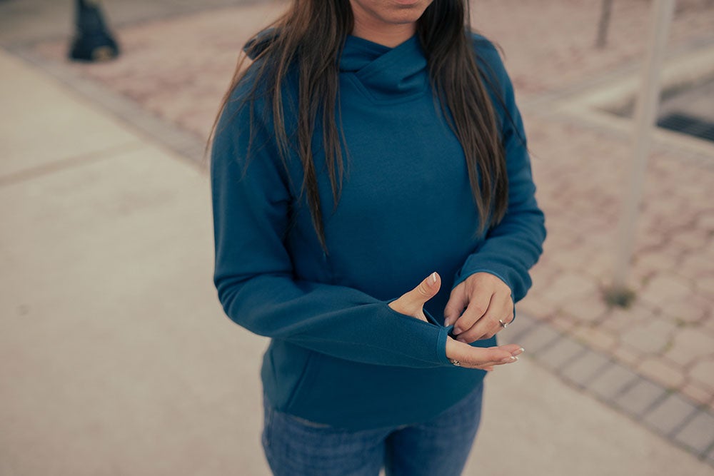 Photography model showcasing a teal hooded sweatshirt with thumbholes and a subtle asymmetrical neckline outdoors against a blurred background of pavement and distant structures