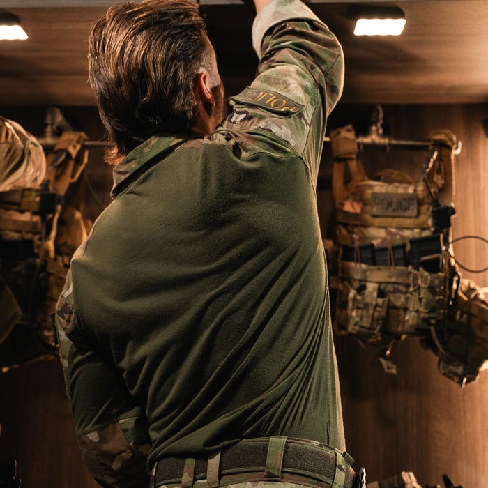 Photograph of a soldier in a dark-green uniform reaching for something in a dimly lit room with camouflage tactical vests hanging on the wall featuring tan and olive green details and POLICE patches visible