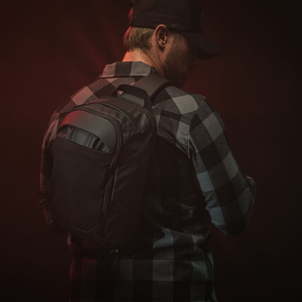 Studio photography featuring a man wearing a black sling backpack with gray and black plaid shirt against a dark red background showcasing the bag's sleek design durable fabric and multiple compartments