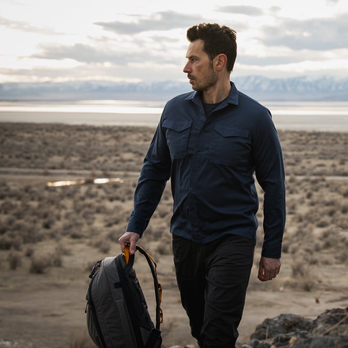 Photography A man in a navy shirt and black pants walks across a desert landscape carrying a gray backpack with orange accents toward snow-capped mountains and a large body of water