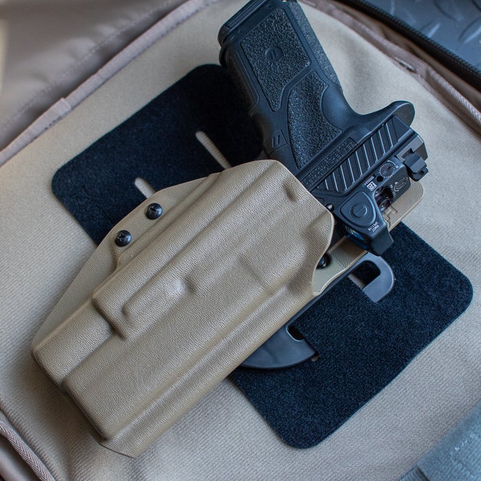 Photograph of a tan polymer handgun holster with a black handgun inside on a tan surface showing two black screws textured polymer and a red button
