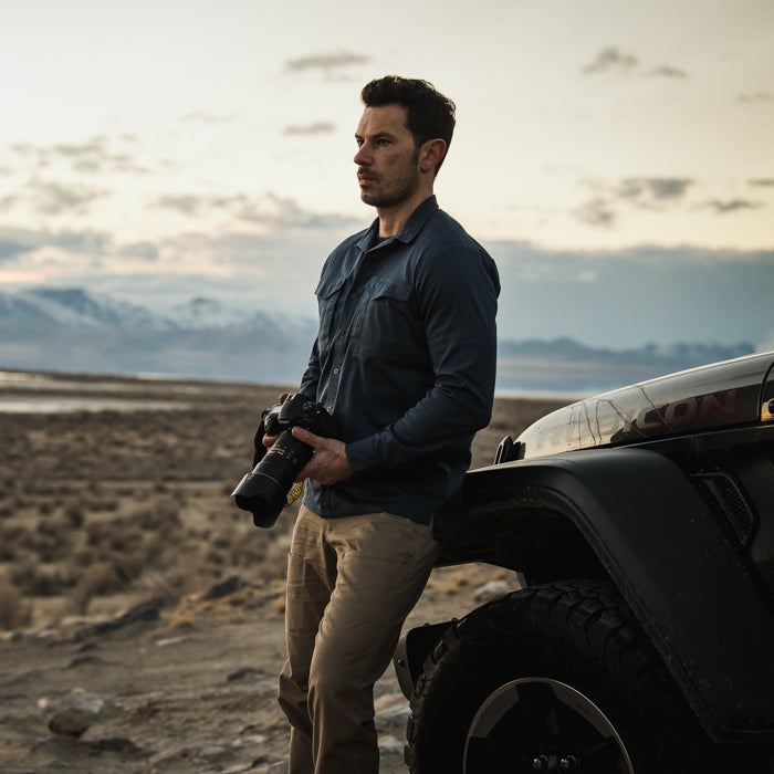 Photograph of a man with a camera leaning against a Jeep in a desert landscape featuring snow-capped mountains and a muted sunset with the photographer wearing khaki pants and a blue shirt