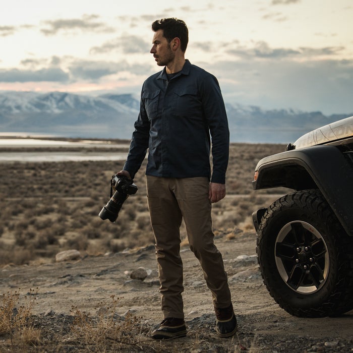 Photograph of a man in a navy shirt and khaki pants holding a camera near a Jeep in a desert landscape featuring snow-capped mountains a dry lakebed and the warm sunset hues