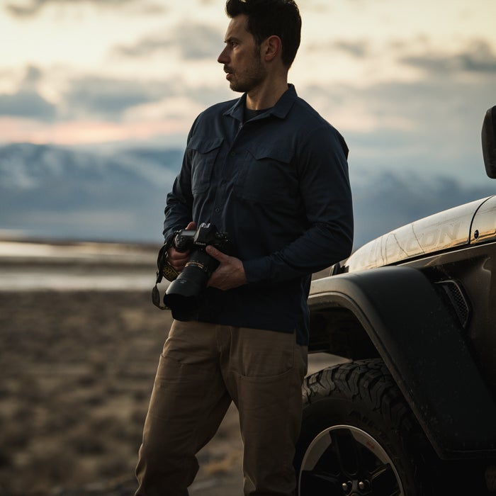 Photography, unknown artist, a man with a Nikon camera stands near a Jeep in a desert landscape featuring distant mountains and a sunset with warm orange and pink hues and a large tire
