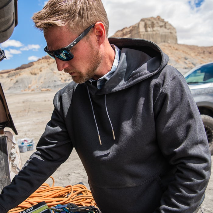 Photograph unknown artist a man wearing a dark gray hoodie with brass drawstring tips near a desert landscape featuring a sandstone rock formation and an orange climbing rope