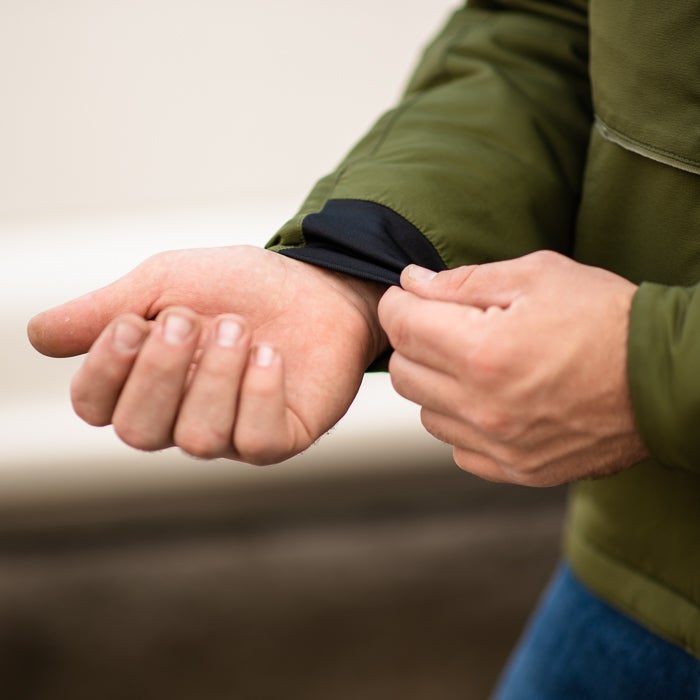 Close-up photograph showing a person adjusting the olive green cuff of a jacket with a black inner lining revealing fair skin and visible fingernails against a blurred background