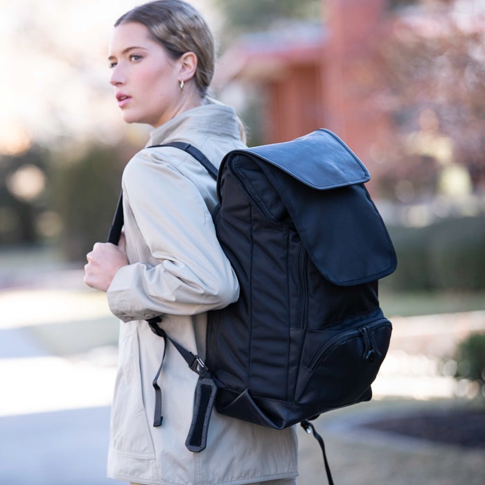 Photograph of a woman wearing a beige jacket and carrying a large black backpack with a flap and buckle detail on a tree-lined street near a reddish-brown house.