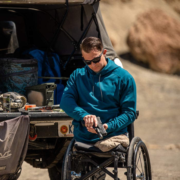 Photograph of a man in a teal hoodie handling a handgun while seated in a wheelchair outdoors near a vehicle with camouflage gear and ammo boxes a sunny day and rocky background