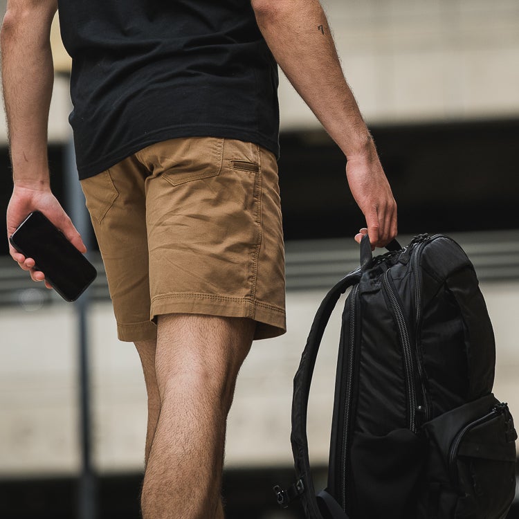 A photograph showing a person wearing tan shorts and carrying a black backpack outdoors near a modern building with a number tattoo and a smartphone