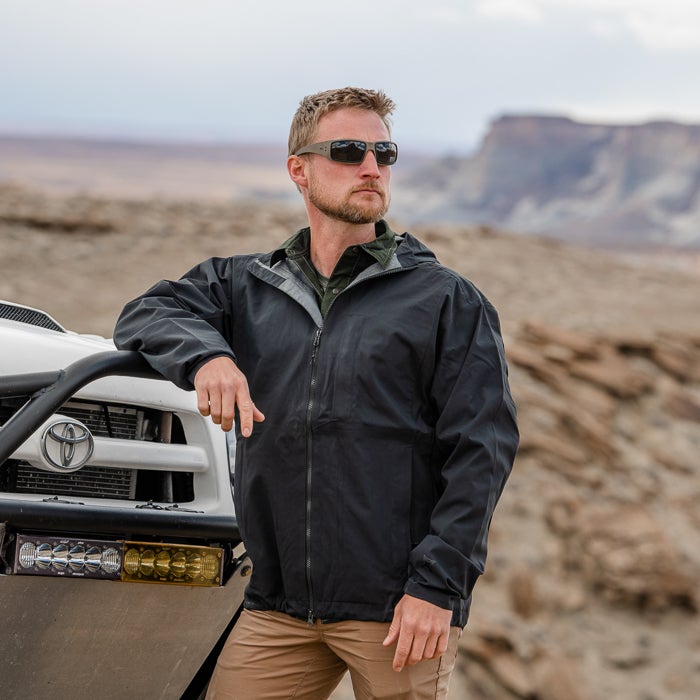 Photograph man wearing black jacket and khaki pants in desert landscape near a Toyota truck with amber light bar featuring rocky mesas and a hazy sky