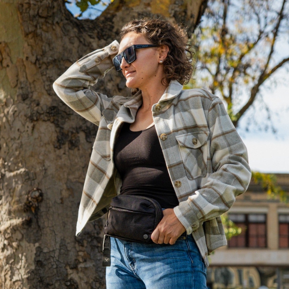 Photograph of a woman wearing a tan and grey plaid shirt, black tank top, blue jeans, and a black fanny pack near a large tree and a vintage-style building with autumn leaves in the background