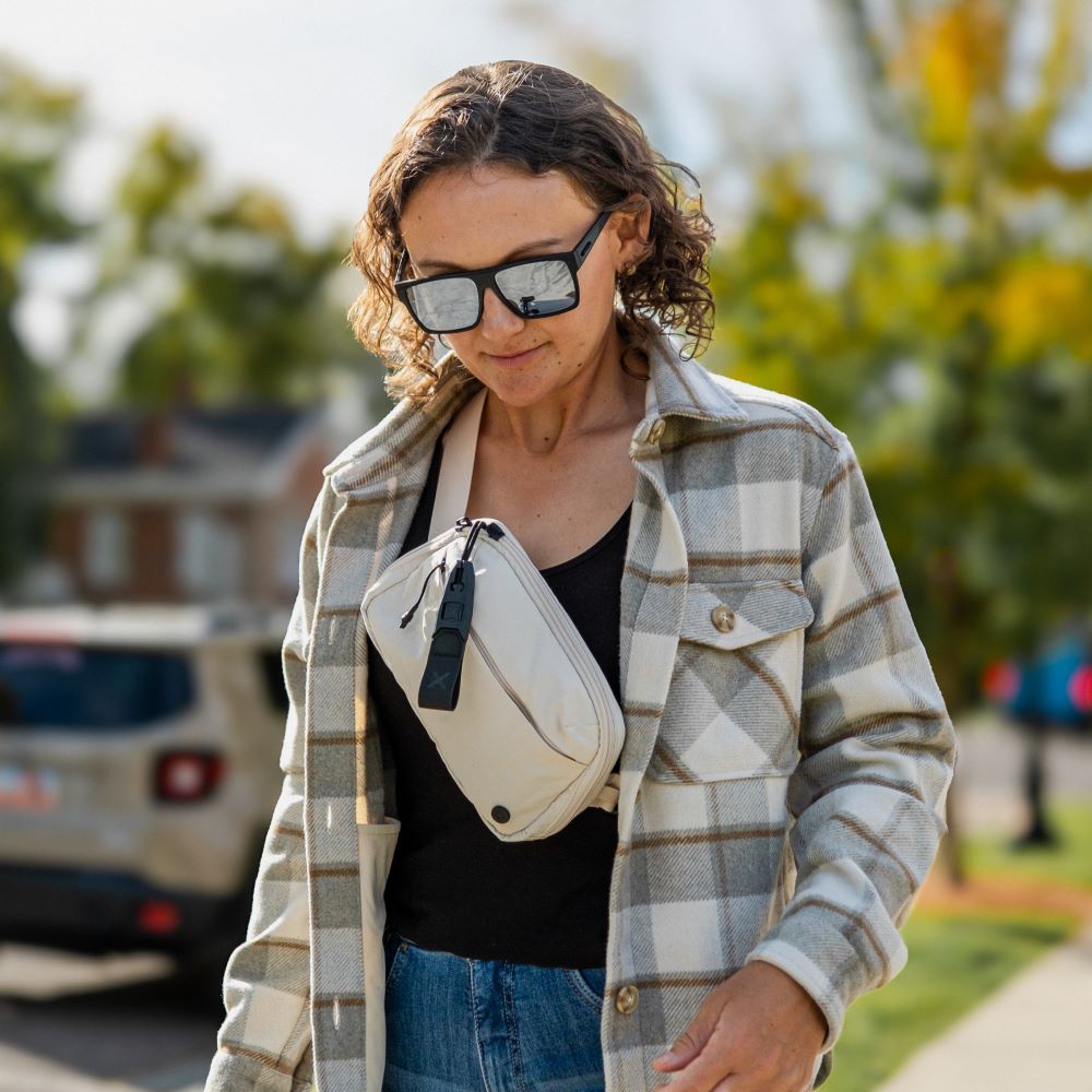 Photograph of a woman wearing a beige crossbody bag and a grey and white plaid shirt walking near a house and trees with reflective sunglasses and a black top