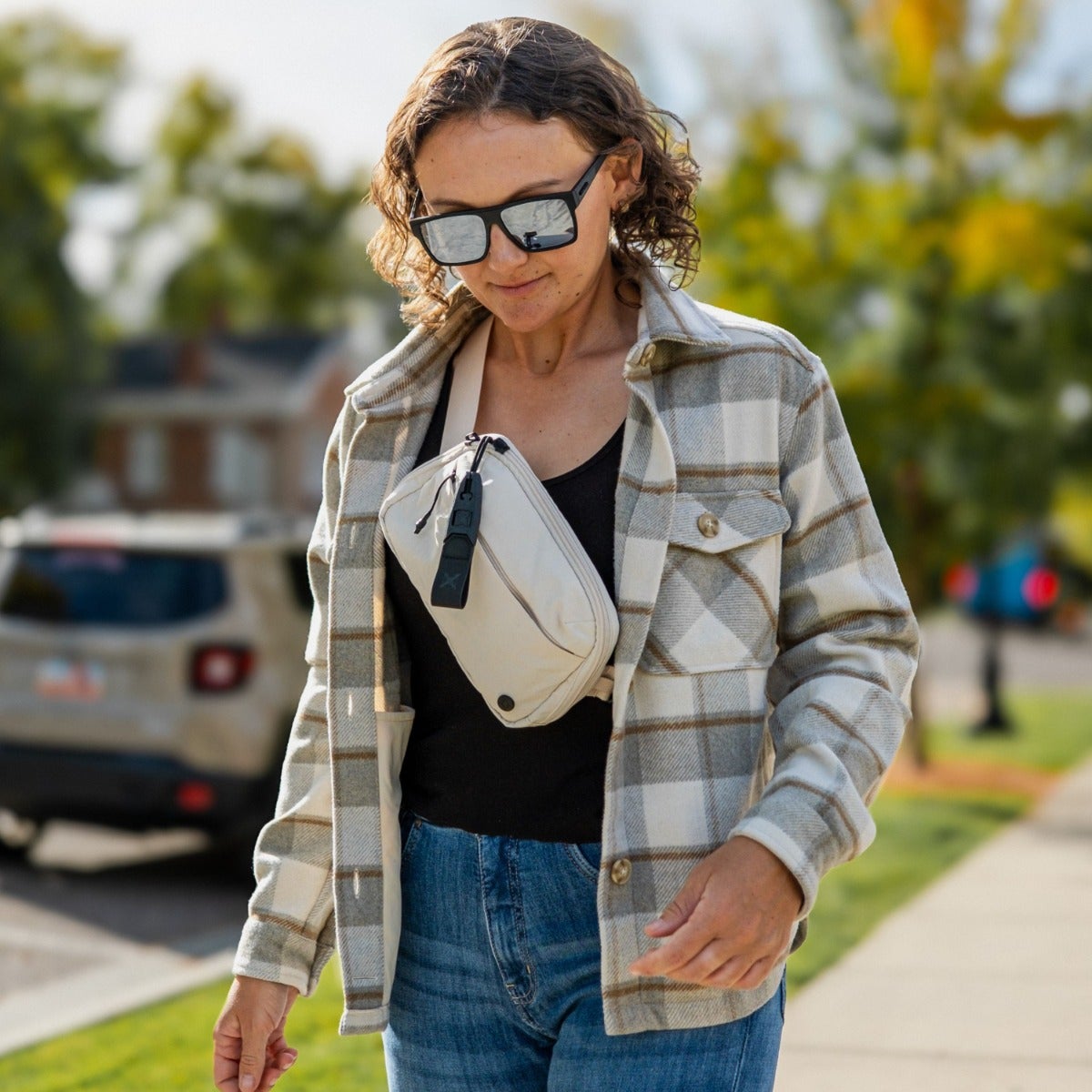 Photograph of a woman wearing a beige crossbody bag and a grey and white plaid shirt walking down a suburban street near a brick house and parked car with reflective sunglasses and a black top