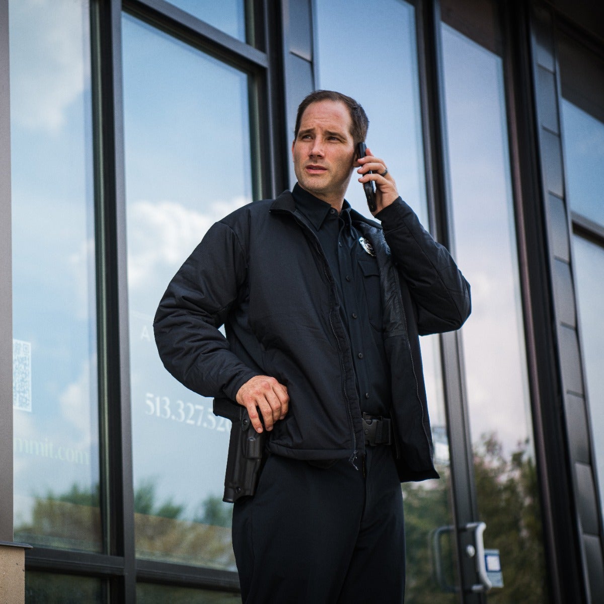 Photograph Security officer on cell phone outside modern building with reflective windows and a partially visible QR code, wearing a black jacket, dark pants, and carrying a sidearm.