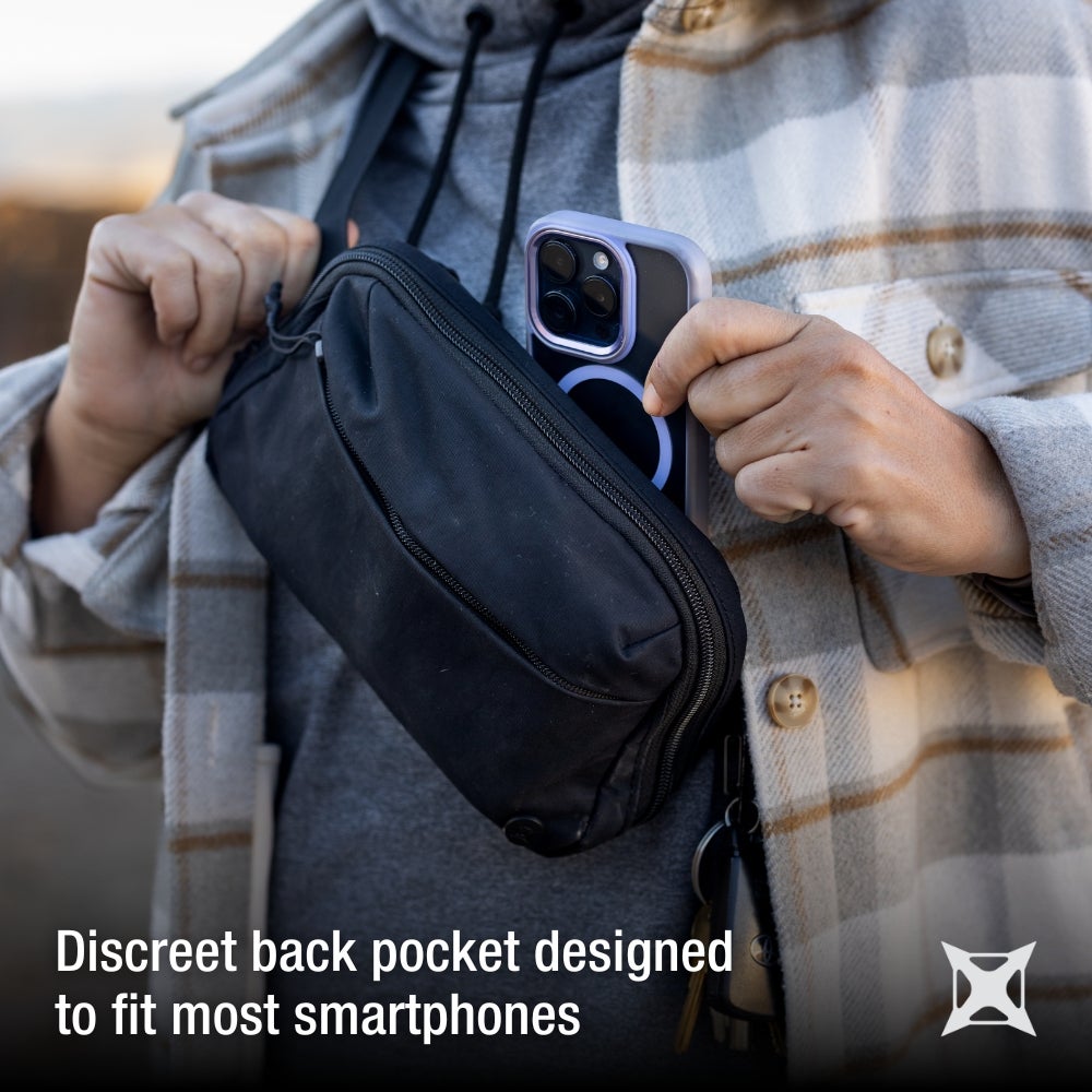 Product photography showing a person placing a smartphone with a lavender case and magnetic ring into a black fanny pack worn across their body the person is wearing a beige and brown plaid shirt and a gray hoodie