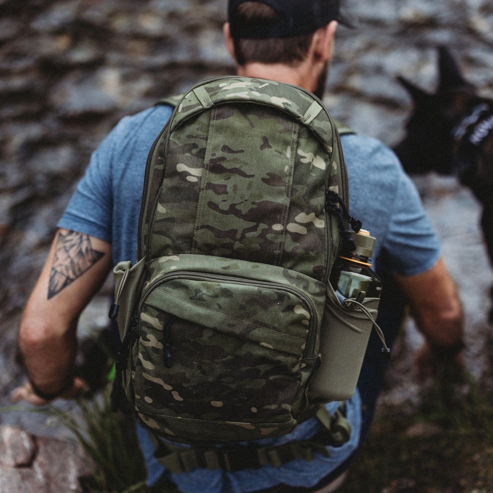 Photograph of a man with a camouflage backpack near a stone wall featuring a geometric tattoo olive green accents and a dark green water bottle