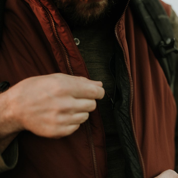 Photograph close-up of a man adjusting a maroon zip-up vest with a dark gray henley undershirt showcasing textured fabric and a copper zipper