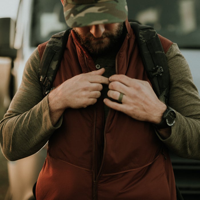 Photograph man wearing a maroon vest olive green shirt and camouflage hat adjusting his vest outdoors near a vehicle a dark green ring and a black watch are visible