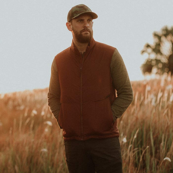 Photograph of a man in a rust-colored vest and camouflage hat standing in a tall grass field with a tree in the background at sunset the vest has zippered pockets and the light creates a warm golden glow