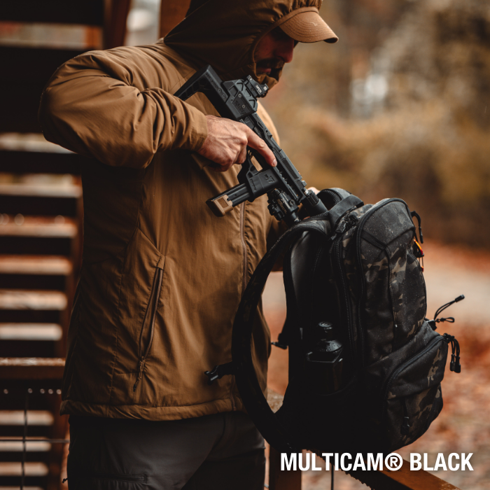 Photography unknown artist a man in a brown jacket stores a black firearm in a Multicam Black backpack near a rustic wooden structure with autumn foliage in the background