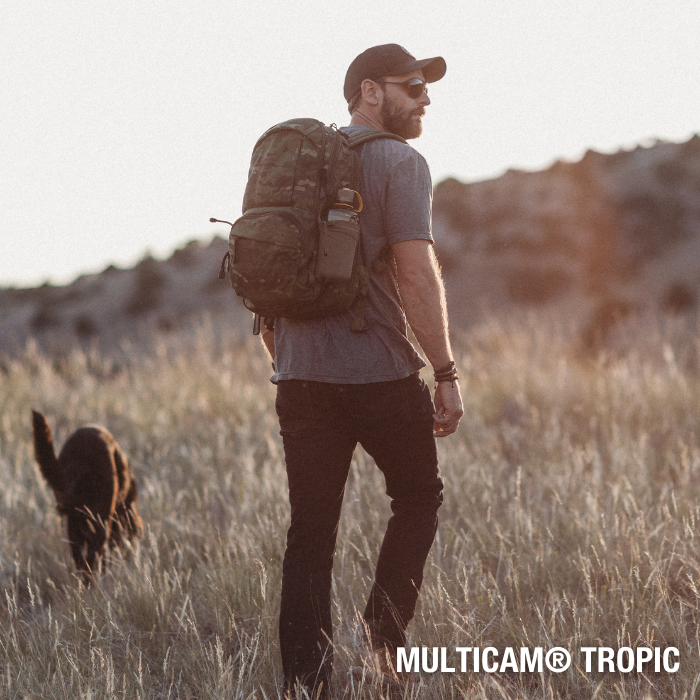Photograph man hiking in a grassy field with his dog a camouflage backpack and a water bottle featuring warm sunset light and earthy tones