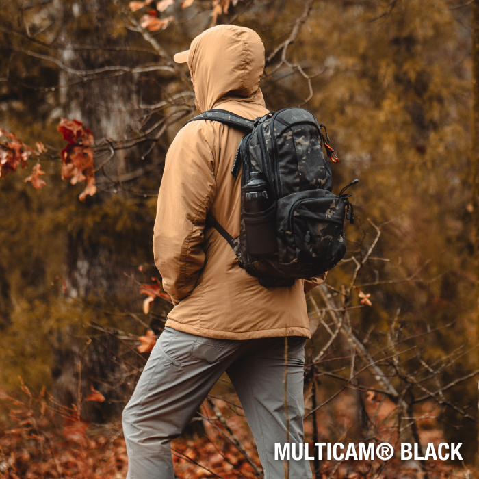 Photography Unknown artist person wearing tan jacket and grey pants with Multicam Black backpack hiking in autumn woods with brown and red leaves and bare branches