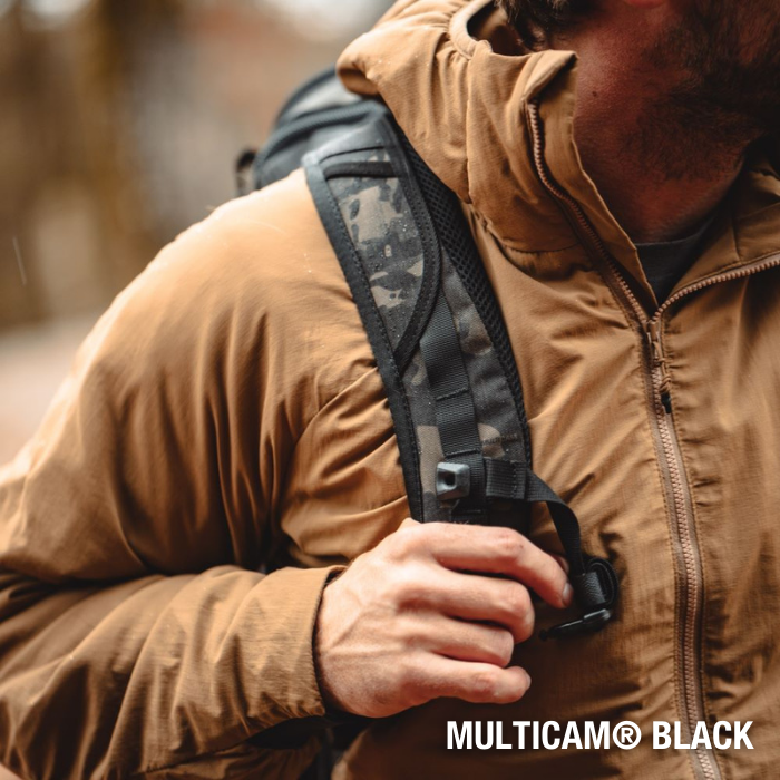 Photograph of a person wearing a tan jacket and Multicam Black backpack outdoors with a dark grey, camouflage strap and a black clasp