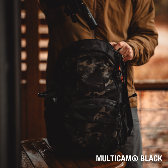 Photograph Multicam Black backpack on wooden table outdoors showing orange zipper pulls and dark camouflage pattern against blurred brown wooden background