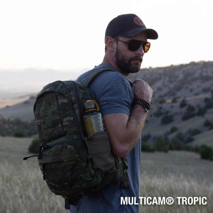 Photography Multicam Tropic backpack model showcasing a man in sunglasses and a baseball cap on a hillside with distant rolling hills and a sunset sky featuring a muted olive green backpack a tan water bottle and a beaded bracelet