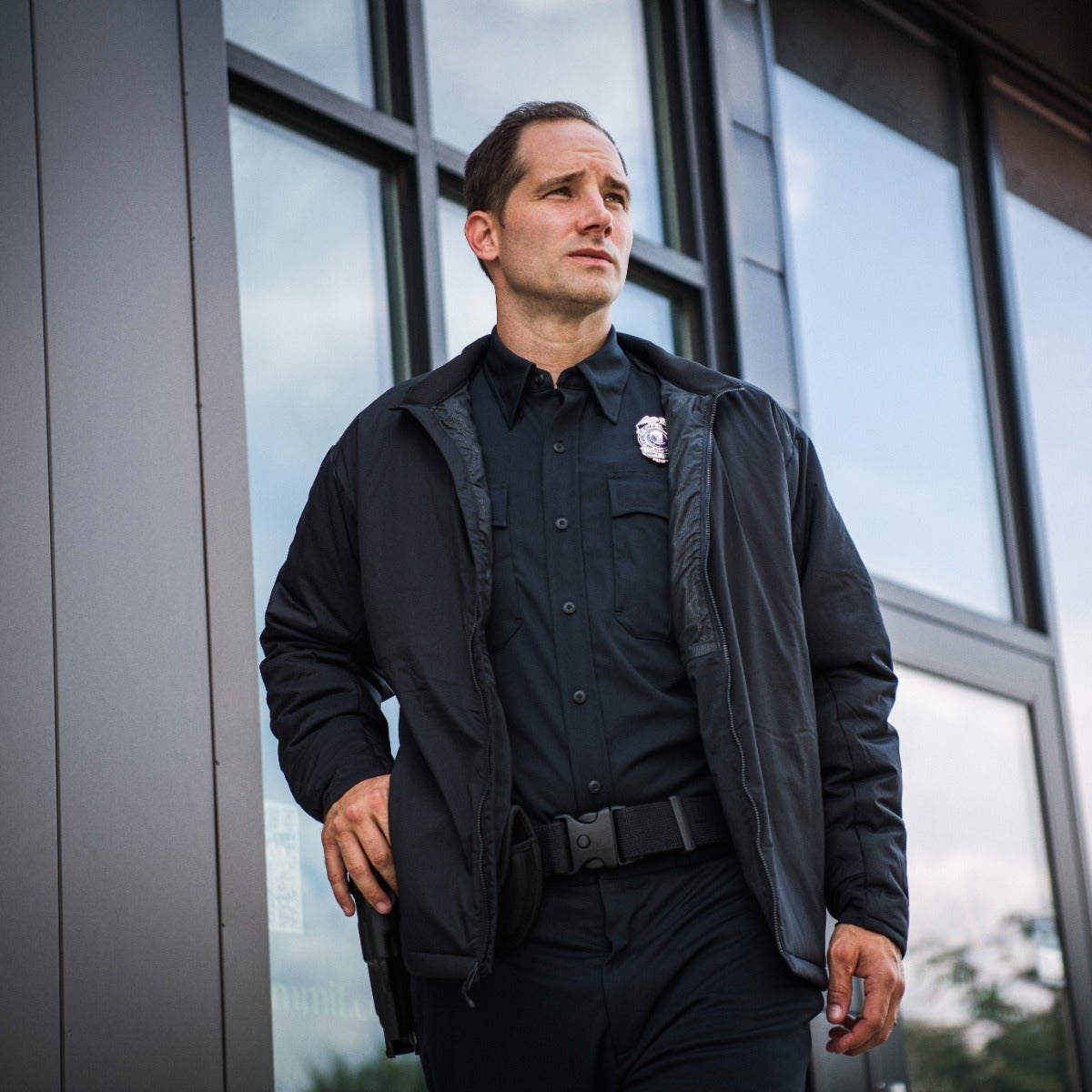 Photograph of a police officer in a black uniform and jacket standing outside a modern building with large glass windows and a dark gray exterior wall the officer's badge, gun, and serious expression are notable details