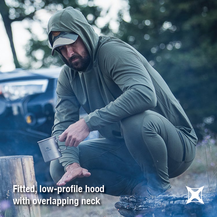 Photography Unknown Artist a man in a green hooded shirt and pants sits near a campfire outdoors next to a vehicle with blue smoke rising and a metallic cup in his hand