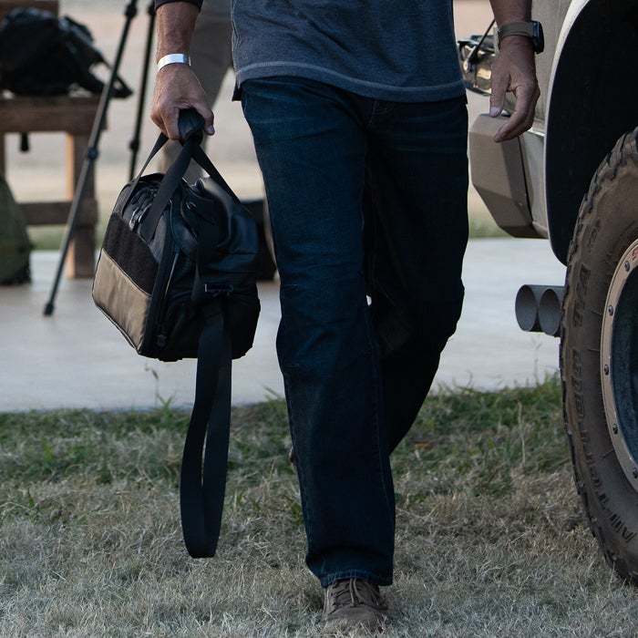 Photograph of a person carrying a black leather bag with a shoulder strap near a vehicle with dual exhaust pipes and a large tire showcasing the bags unique stitching and a silver bracelet on their wrist