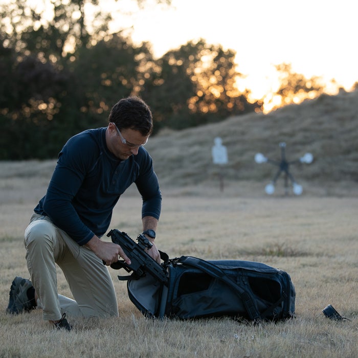 Photograph man kneeling outdoors near shooting range targets packing a black firearm into a dark grey backpack wearing khaki pants and a navy shirt with a watch and safety glasses