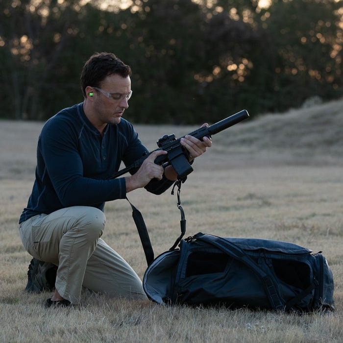 Photograph man kneeling in a field with a suppressed rifle and tactical backpack wearing safety glasses and earplugs at sunset
