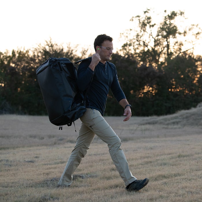 Photograph of a man carrying a large black duffel bag across a field at sunset wearing khaki pants, a dark shirt, safety glasses, and earplugs, featuring trees in the background.