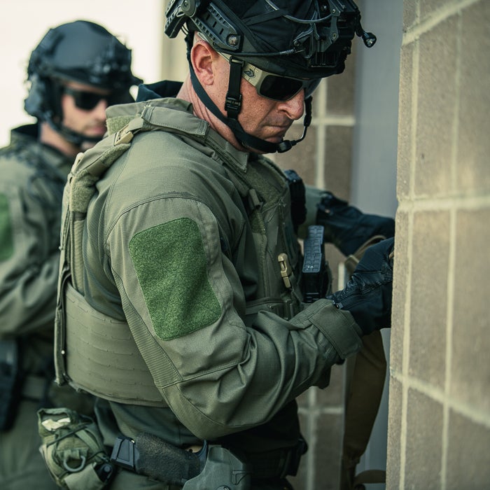 Photograph of a SWAT officer in olive green tactical gear and ballistic vest next to a brick wall with a colleague in the background featuring a dark helmet, tactical gloves and a green arm patch