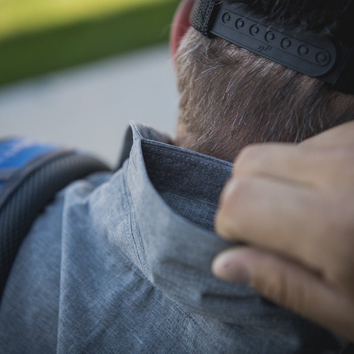 Close-up photograph showing a man adjusting the gray heathered collar of his shirt with a subtle textured fabric and dark stitching detail near the nape of his neck his graying hair is visible under a black hat