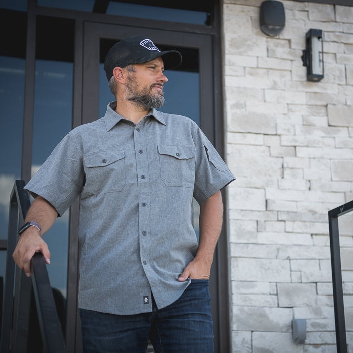 Photography Unknown A man in a grey shirt and black cap stands on steps outside a building with a light brick exterior and dark modern door a watch and a stylish beard are visible