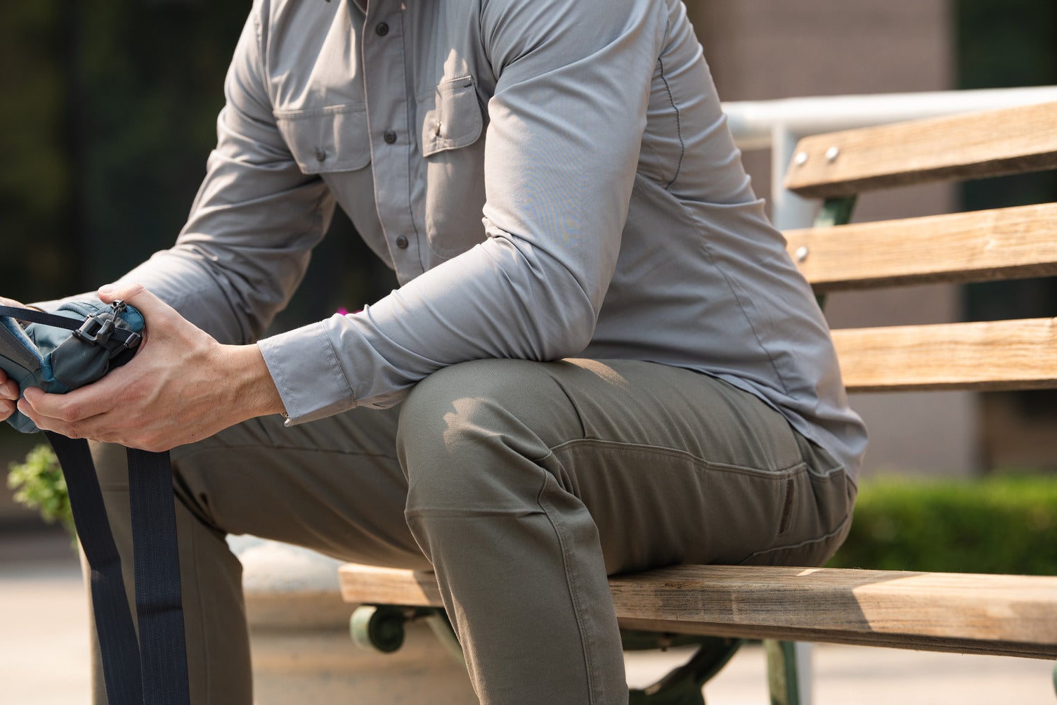 Photograph A man in a grey shirt and olive green pants sits on a weathered wooden bench holding a dark blue bag with black straps outdoors near green plants