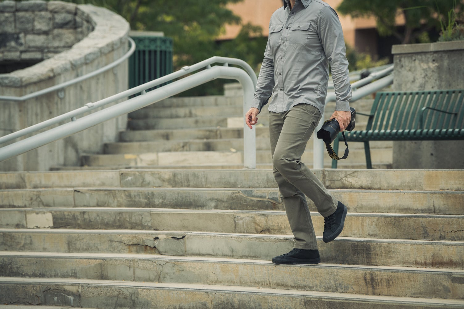 Photograph of a man in olive pants and a grey shirt descending a concrete staircase with a metal railing and a green bench near a stone wall carrying a professional camera showcasing the man's black shoes and the camera's yellow strap