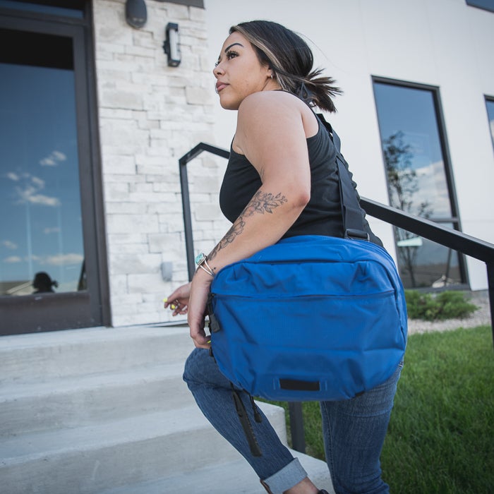 Photograph a woman with a vibrant blue bag ascending steps outside a modern building featuring light stone and large windows a floral arm tattoo and turquoise jewelry are visible