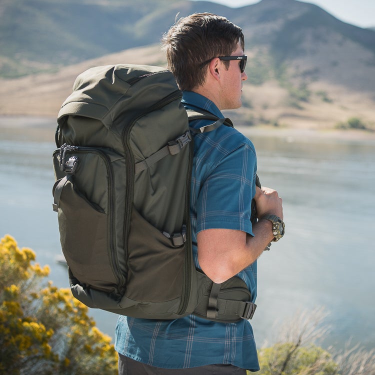Photograph of a man wearing a large olive-green backpack with multiple pockets and compression straps by a lake near mountains showcasing yellow wildflowers and his blue plaid shirt and smartwatch